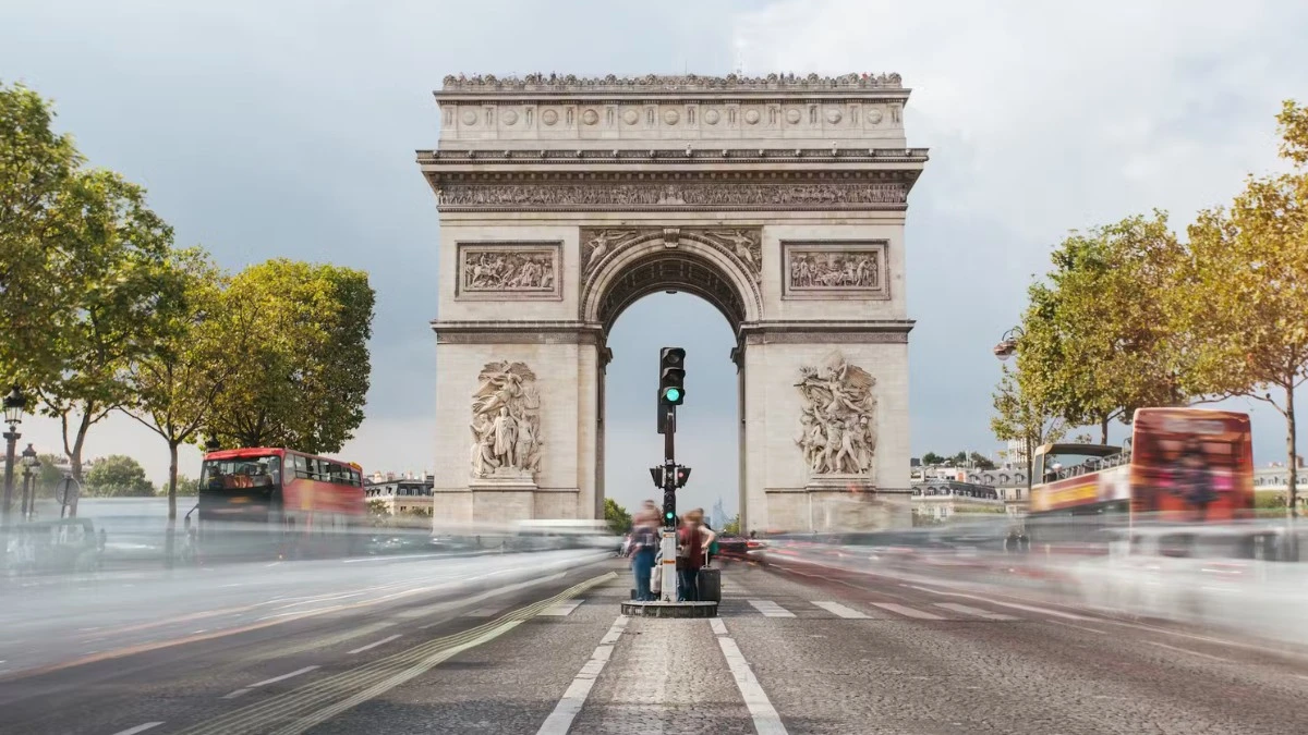 The Arc de Triomphe with blurred traffic lights and bus trails, indicating movement, and trees on either side.