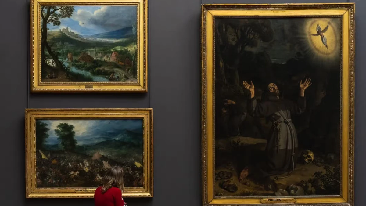 A person viewing paintings displayed on a wall inside the Louvre Museum.