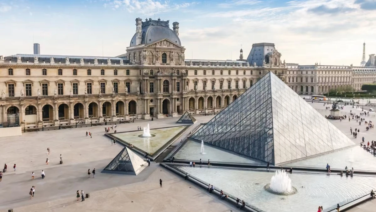 An aerial view of the Louvre Museum complex with its pyramids and fountains under a cloudy sky.
