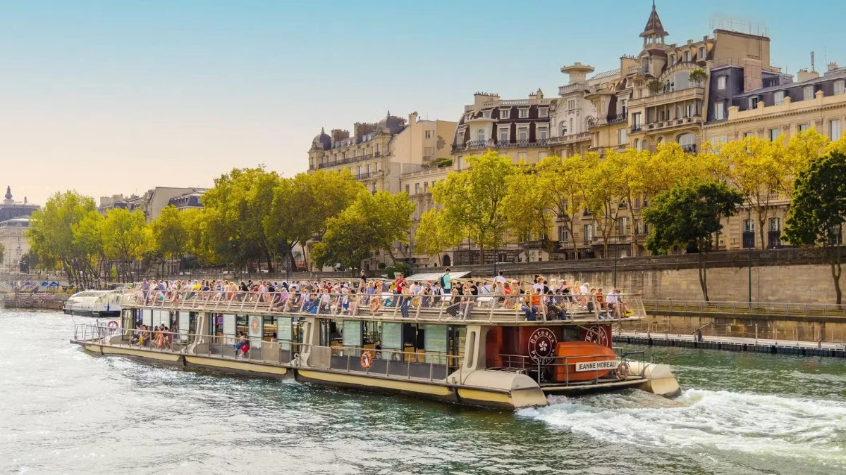 A crowded tour boat moving along the Seine River with classic Parisian buildings and trees lining the bank.