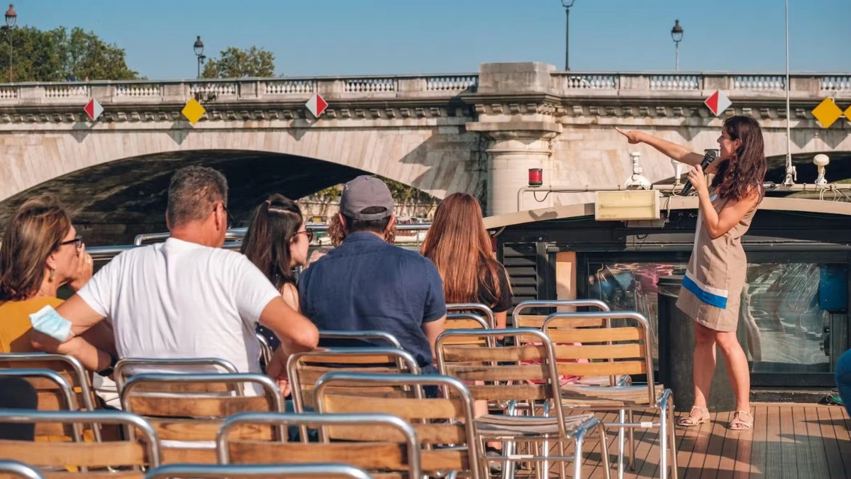 A tour guide speaking to passengers on a boat on the Seine River, with a stone bridge in the background.