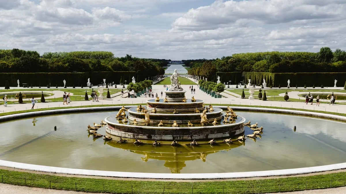 A wide view of the Gardens of Versailles with a large circular fountain in the foreground and a long canal in the distance.